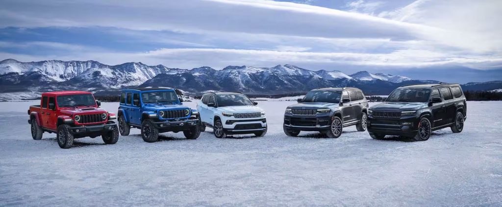 Five Jeep SUVs and trucks parked on a snowy landscape with mountains in the background.